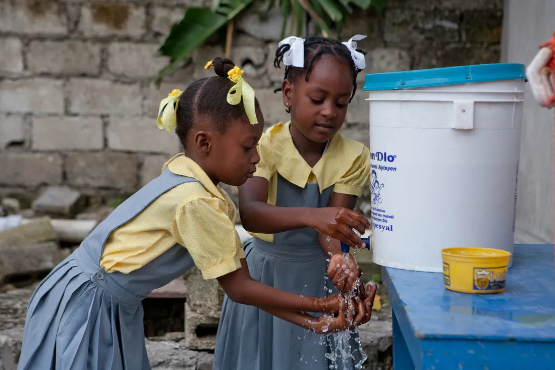 Two young Haitian girls wearing denim dresses over yellow shirts wash their hands at a station.