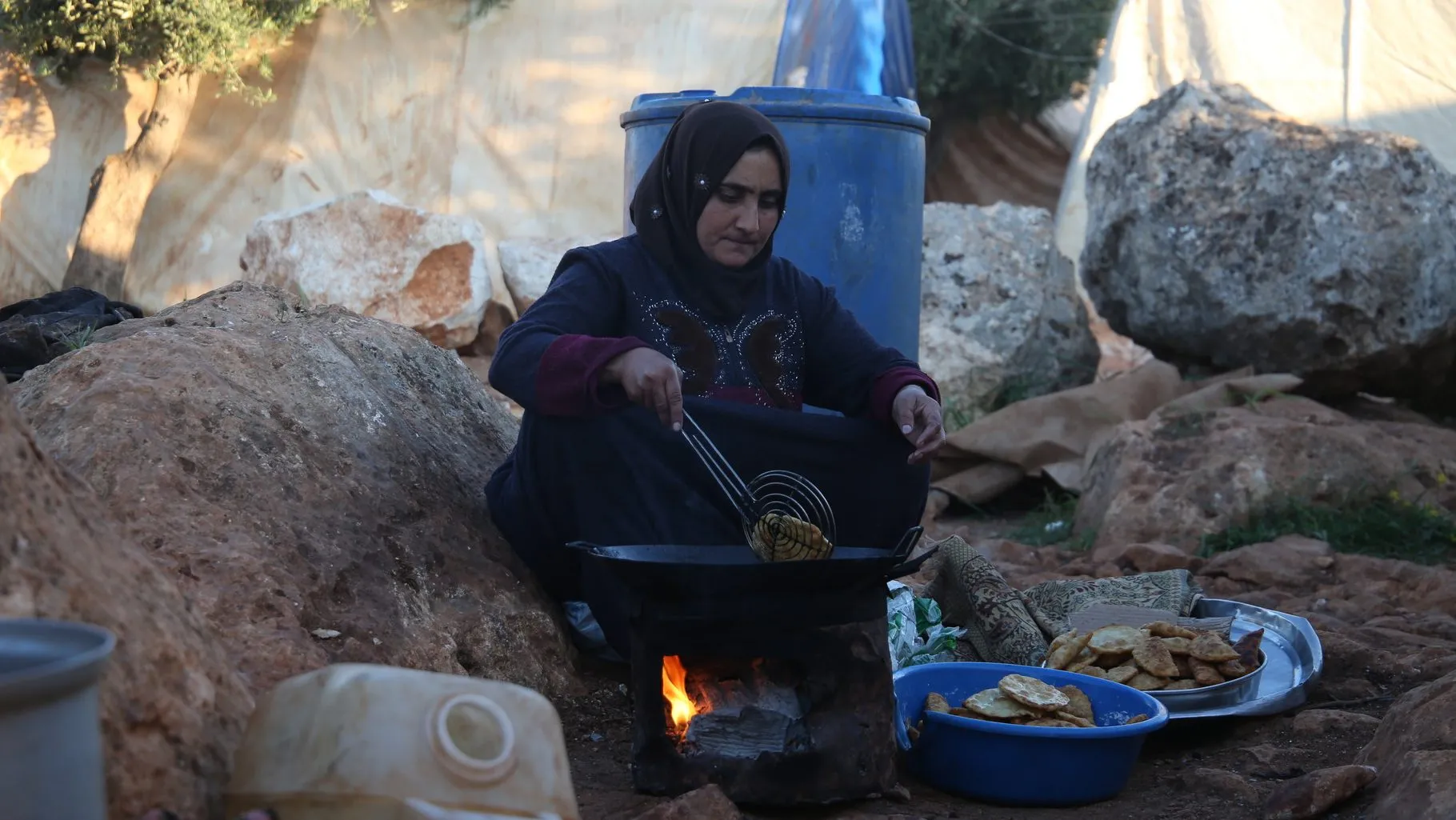 A Syrian woman in dark clothing cooks over an open fire in a displacement camp